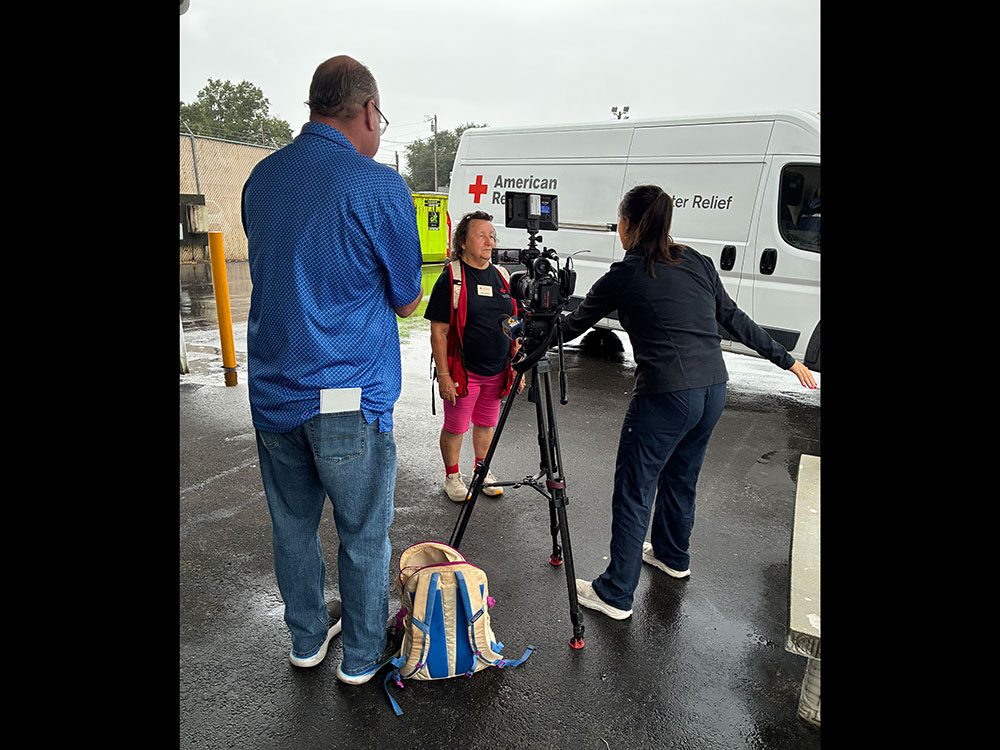 Red Cross volunteer Helen Miller in front of a news camera with two other people in front of her and a Red Cross van in the background.