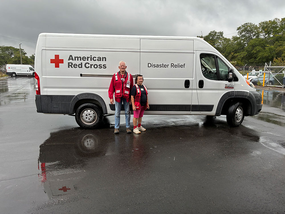 Red Cross volunteer Helen Miller and Dan Wood stand in front of their emergency response vehicle in a parking lot on a cloudy rainy day.