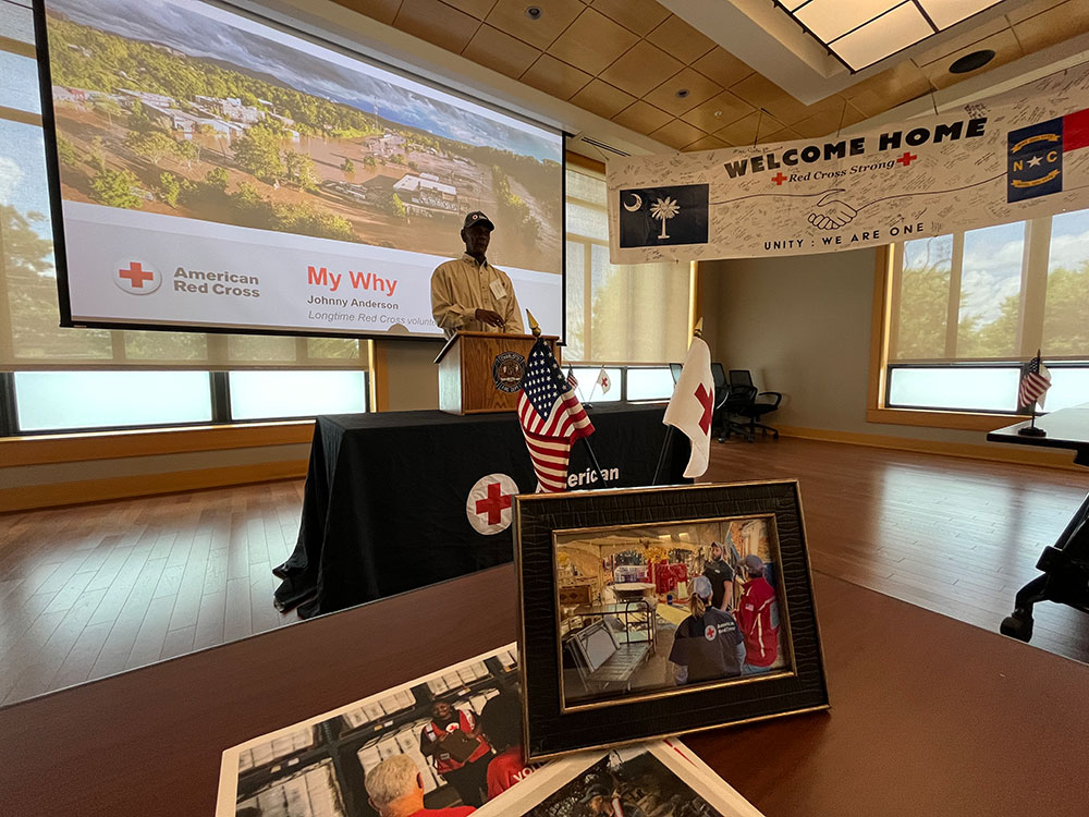 Jonny Anderson standing at podium in a room with welcome home banner, a projection screen, desk with picture, small american flag and red cross flag.