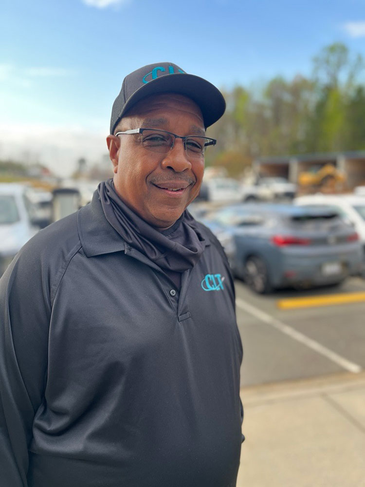 Reginald LaMon in a black cap and jacket with a blue logo stands in a parking lot. He smiles warmly on a sunny day, with blurred cars and trees in the background.