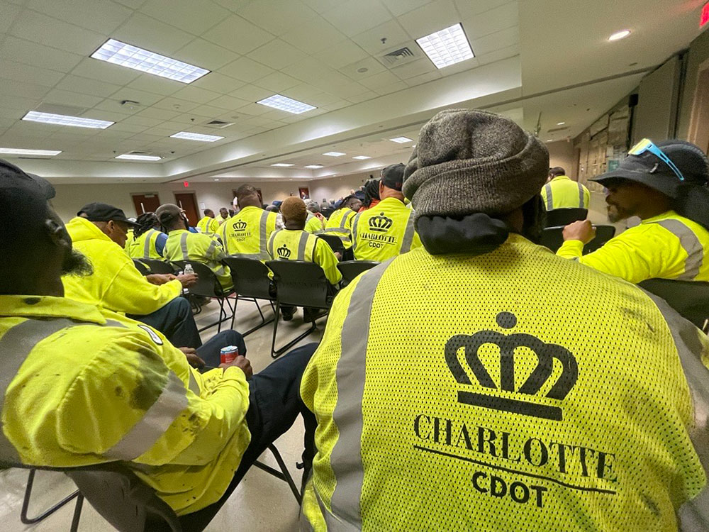 Charlotte DOT employees in bright yellow safety vests with "Charlotte CDOT" logos sit in rows during an indoor meeting.