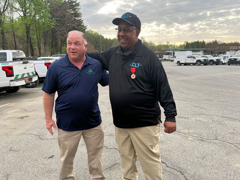 Reginald LaMon stands next to friend and former colleague 'Billy' in a parking lot, smiling. One wears a blue polo, the other a black shirt with "CLT" and a red medal. Parked trucks are visible behind them.