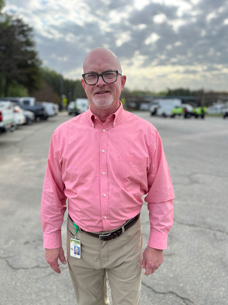Steve Whitten in a pink shirt stands in a parking lot, smiling slightly. The background shows parked cars and a cloudy sky.
