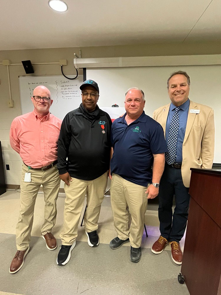 Steve Whitten, Reginald Lamon, 'Billy', and Jason Gudzunas stand side by side in a classroom. They are all smiling, with a whiteboard in the background.