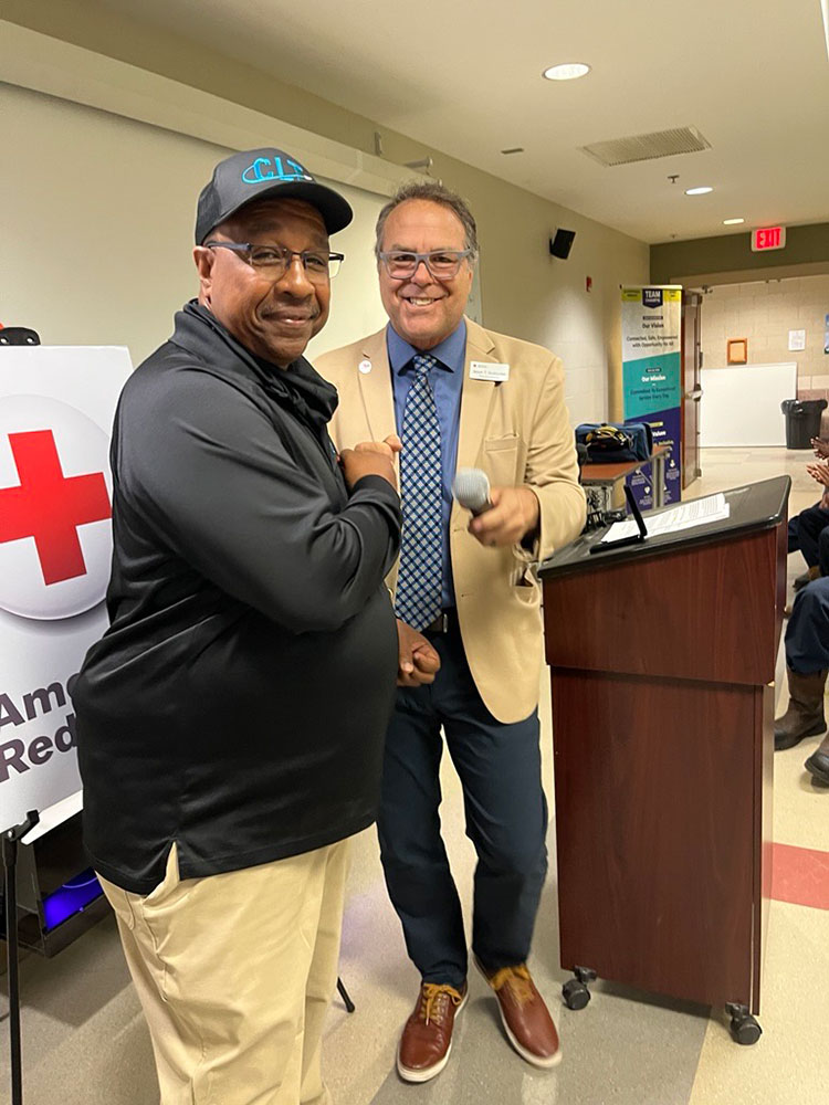 Reginald LaMon and Jason Gudzunas smiling and shaking hands in front of a podium with a microphone. One wears a suit and tie, the other a cap and casual jacket. Red Cross banner visible.
