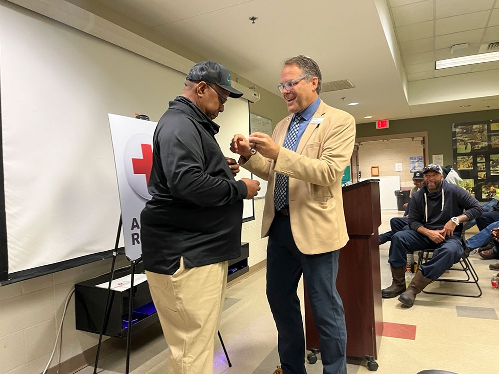 Jason Gudzunas in a tan blazer awards a pin to Reginald LaMon wearing a cap in a room with an audience. A Red Cross poster is visible on a stand.