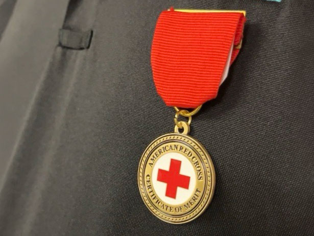 A gold medal with a red cross in the center, labeled "American Red Cross Certificate of Merit," hangs on a red ribbon against a dark fabric background.