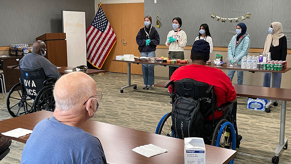 Red Cross HS Club members speaking to room of veterans in wheelchairs.