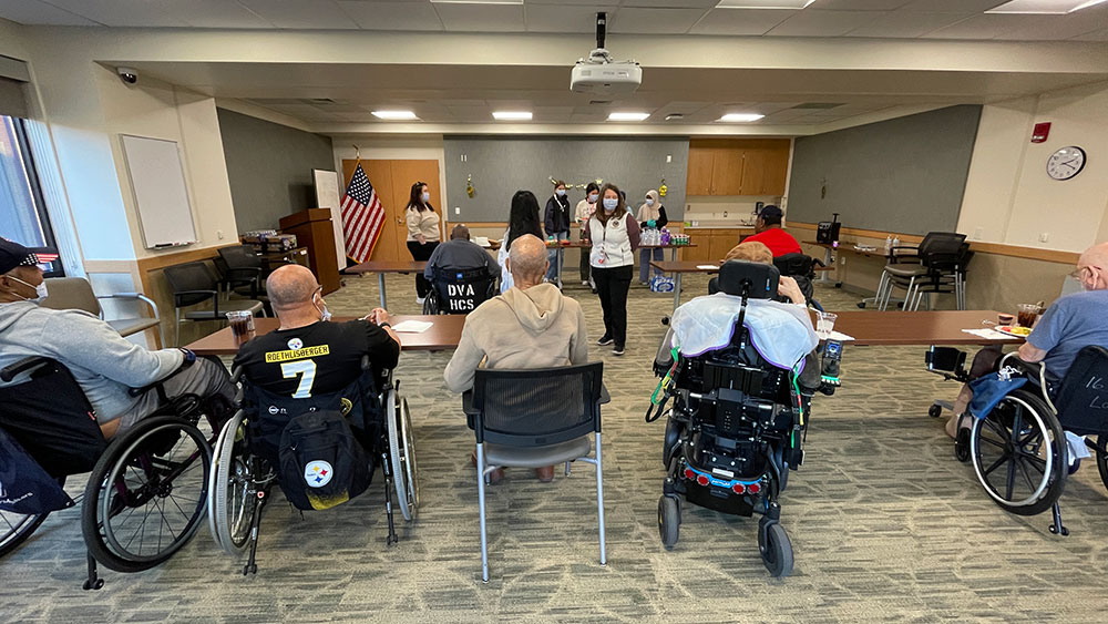 Red Cross HS Club members standing behind tables while a member speaks to room of veterans in wheelchairs.