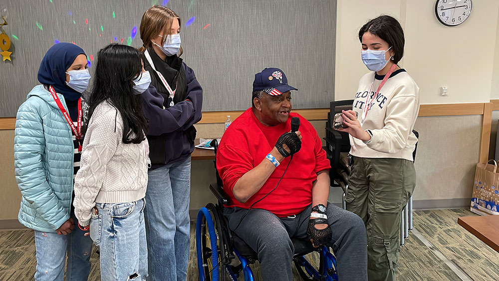 Red Cross HS members wearing masks stand next to veteran in wheelchair speaking using a microphone.