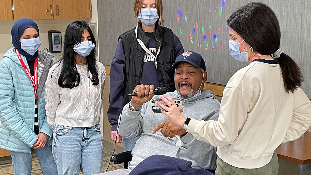 Red Cross HS members wearing masks with one member holding a smartphone and showing it to a veteran in wheelchair who is holding a microphone.