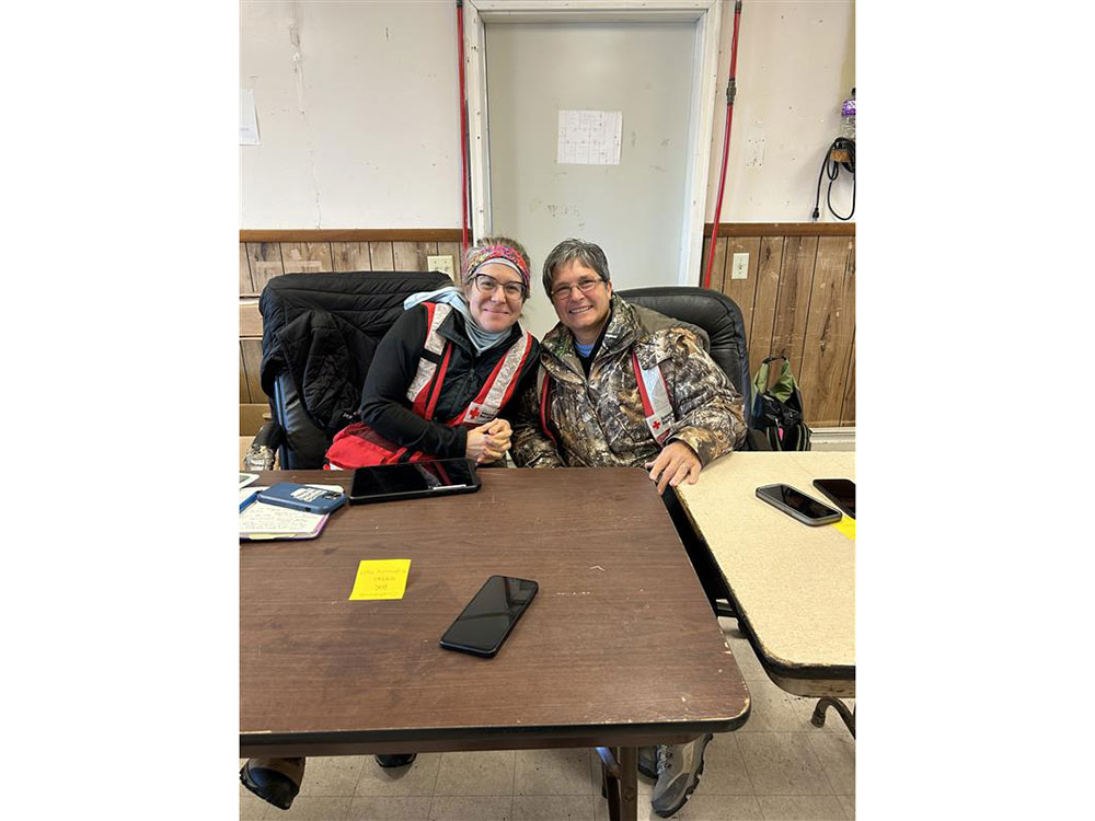 Two Red Cross volunteers wearing jackets and sitting at a table in a room.