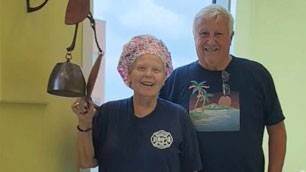 Sherry Kemp smiles and rings a bell in a hospital hallway, wearing a floral cap, with a man beside her smiling.