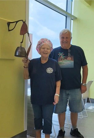Sherry Kemp smiles and rings a bell in a hospital hallway, wearing a floral cap, with a man beside her smiling.