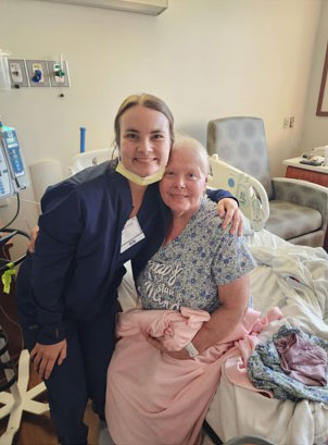 A smiling healthcare worker in scrubs and Sherry Kemp hug beside a hospital bed.