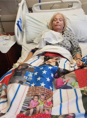 Sherry Kemp resting in a hospital bed, covered by a quilt with family photos and stars.