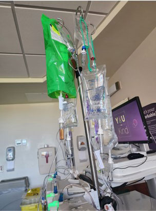 A hospital room showing multiple IV bags and tubes on a metal stand. A monitor displaying "Stay Kind" is visible.