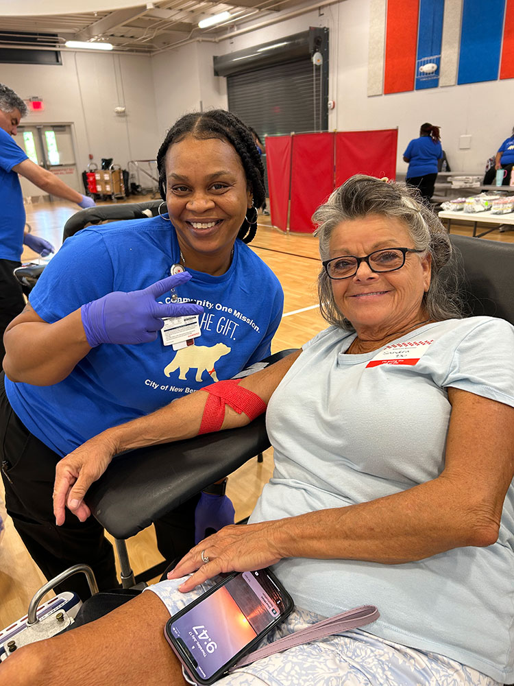 Sandra Ammons donating blood with a red cross nurse standing next to her and giving a peace sign.