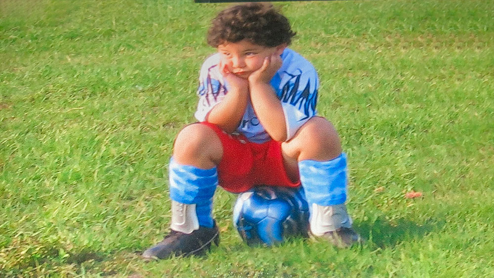 Daniel Agnelli in a soccer uniform, sits on a soccer ball on a grassy field.