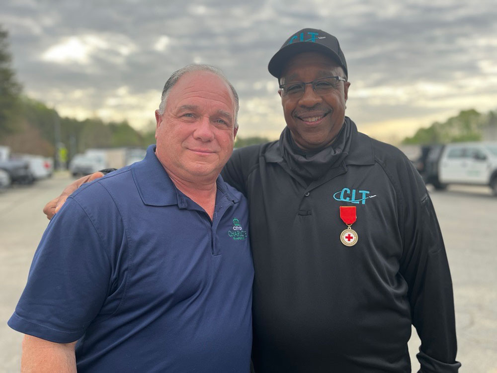 Reginald Lamon stands next to 'Billy' with the Charlotte DOT, smiling and embracing outdoors. One wears a blue shirt, the other a black jacket with a badge. Overcast sky and parked vehicles in the background.