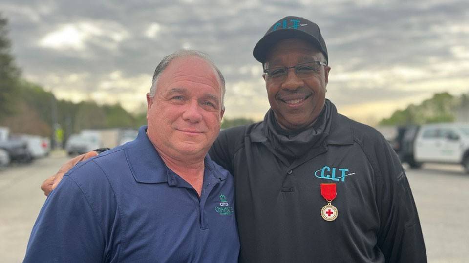 Reginald Lamon stands next to 'Billy' with the Charlotte DOT, smiling and embracing outdoors. One wears a blue shirt, the other a black jacket with a badge. Overcast sky and parked vehicles in the background.