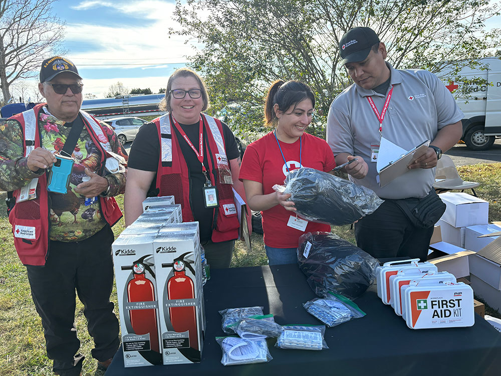 Red Cross volunteers standing next to a table with fire extinguishers, first aid kits and other items.