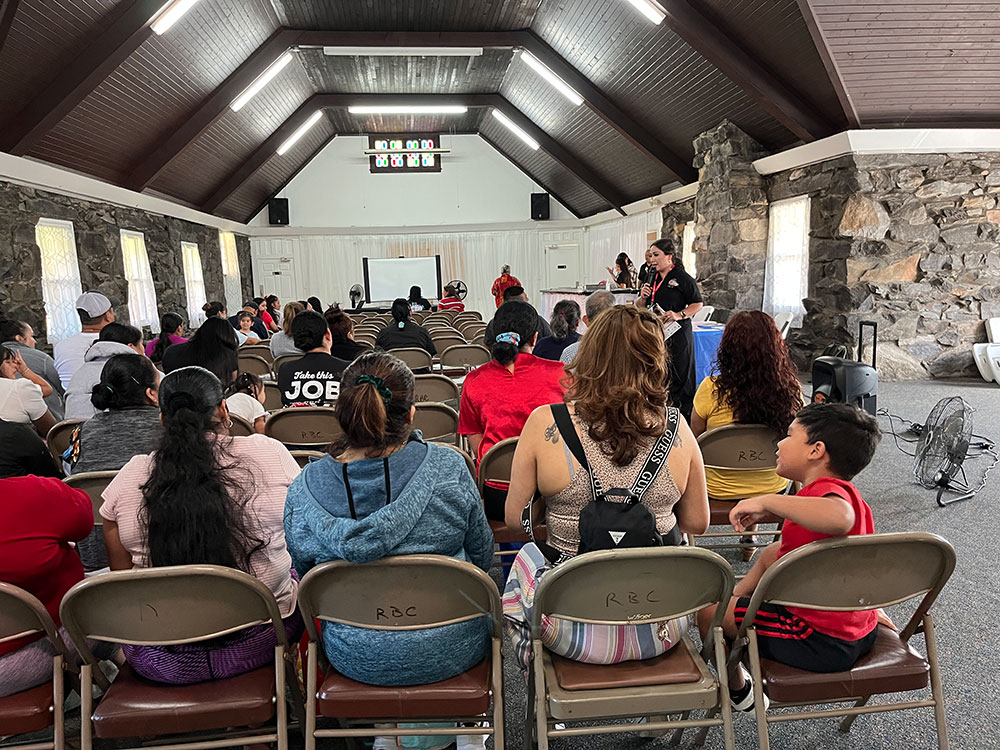 room full of people sitting in folding chairs while a person speaks on a mic.