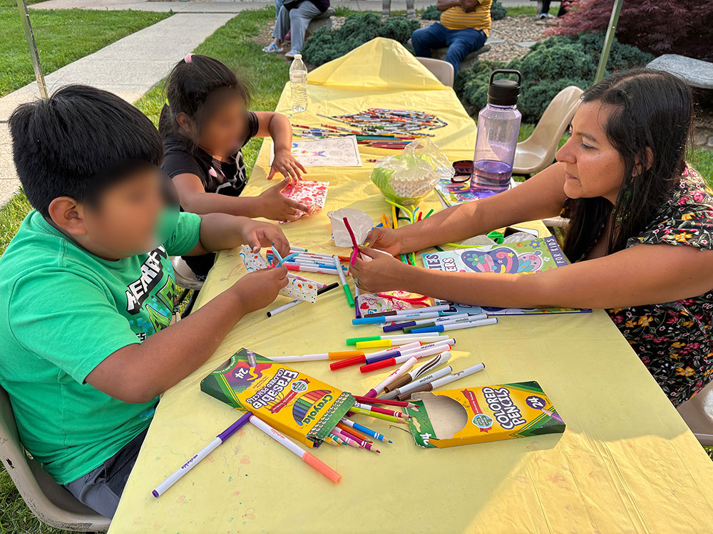 children sitting at table using crayon pencils and markers with an adult helping them.