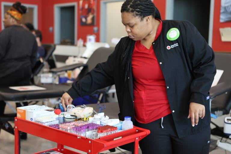 A person in a red shirt and red cross jacket standing in front of a red cart with medical supplies