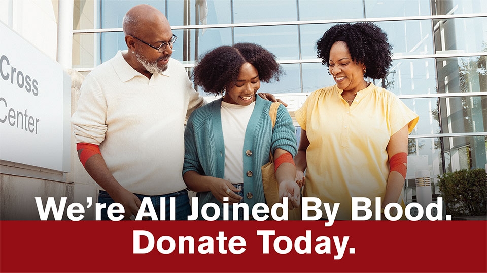 A group of three diverse people smiling outside a building labeled Red Cross Blood Donation Center, with bandages on their arms. Text: "We're All Joined By Blood. Donate Today."