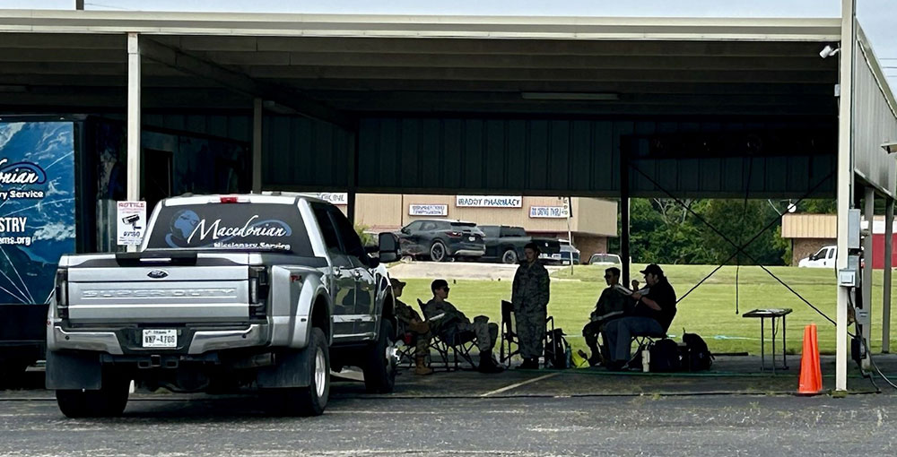A covered outdoor area with people sitting on folding chairs, gathered in the shade beside a parked truck labeled "Macedonian Missionary Service."