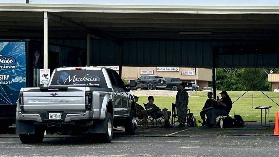 A covered outdoor area with people sitting on folding chairs, gathered in the shade beside a parked truck labeled "Macedonian Missionary Service."