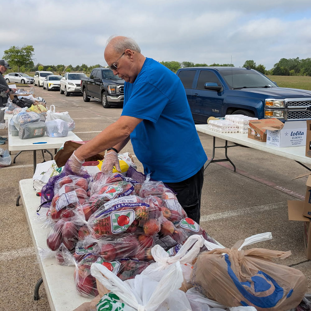 Larry Mueller sorts bagged produce at an outdoor food distribution event. Tables with goods and parked cars are visible under a cloudy sky.