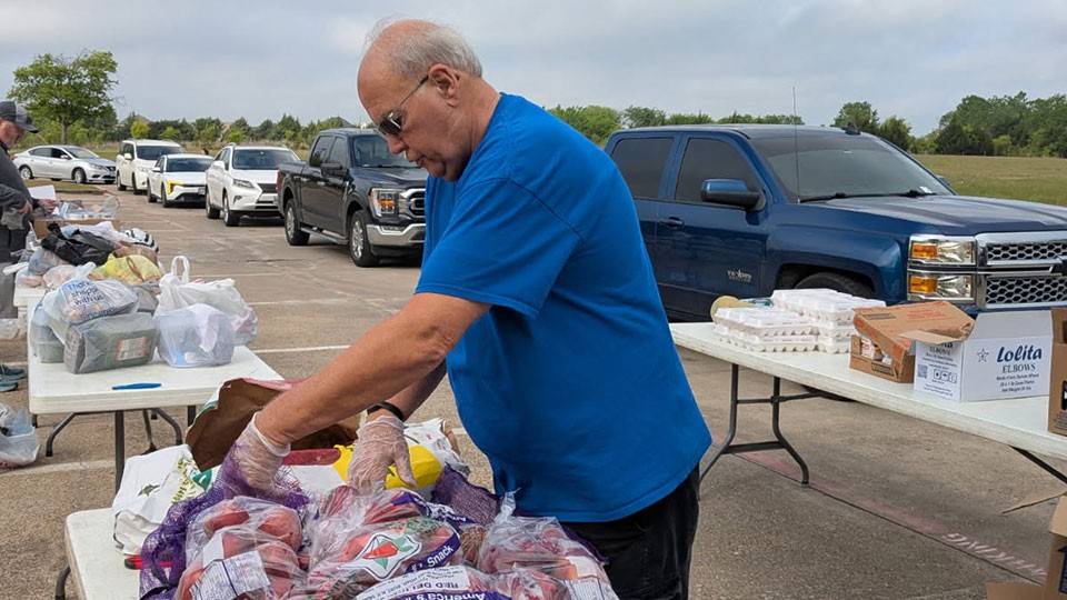 Larry Mueller sorts bagged produce at an outdoor food distribution event. Tables with goods and parked cars are visible under a cloudy sky.
