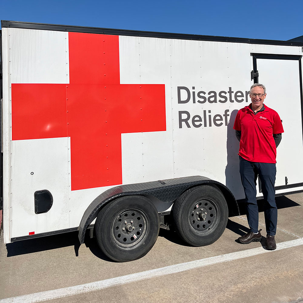 Mark Fitzgibbons in a Red Cross shirt stands smiling beside a disaster relief trailer with a large red cross symbol, under a clear blue sky.