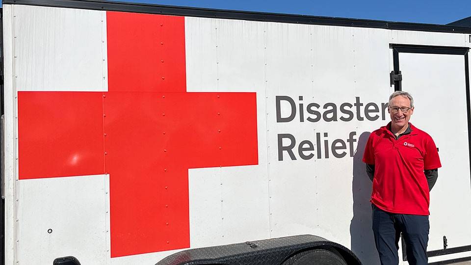 Mark Fitzgibbons in a Red Cross shirt stands smiling beside a disaster relief trailer with a large red cross symbol, under a clear blue sky.