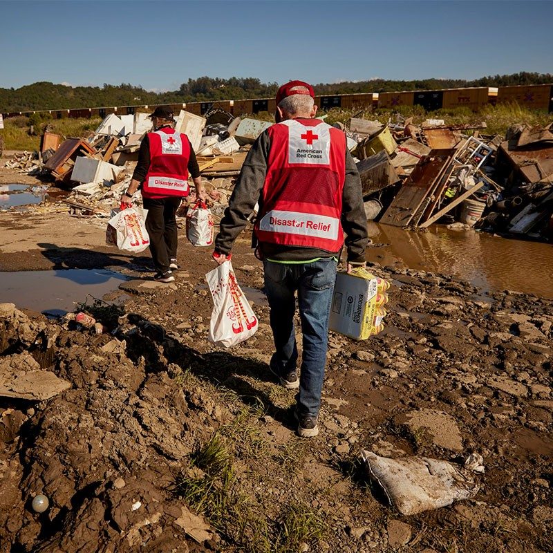 Two Red Cross volunteers carrying supplies across damaged area.