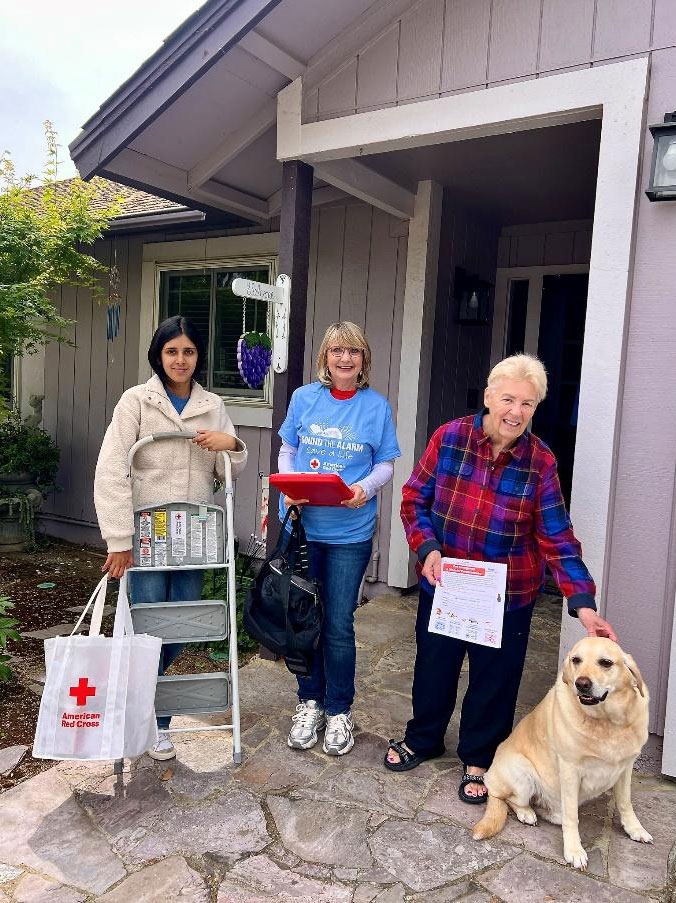 Three women stand outside a house. One holds a ladder and Red Cross bag, another holds a clipboard, and an older woman pats a yellow Labrador.