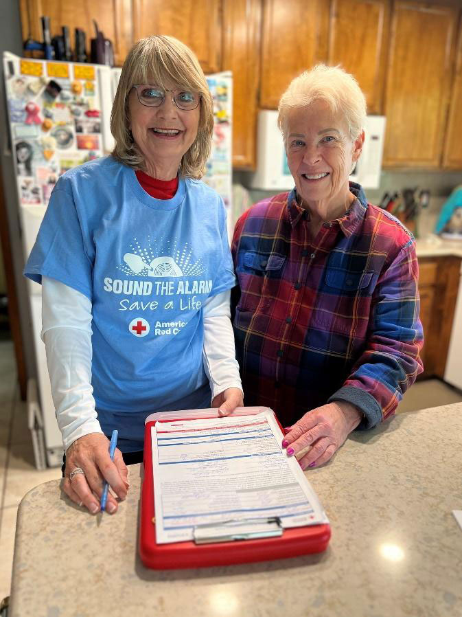 Two smiling women stand in a kitchen. One wears a "Sound the Alarm" shirt, holding a clipboard with forms.
