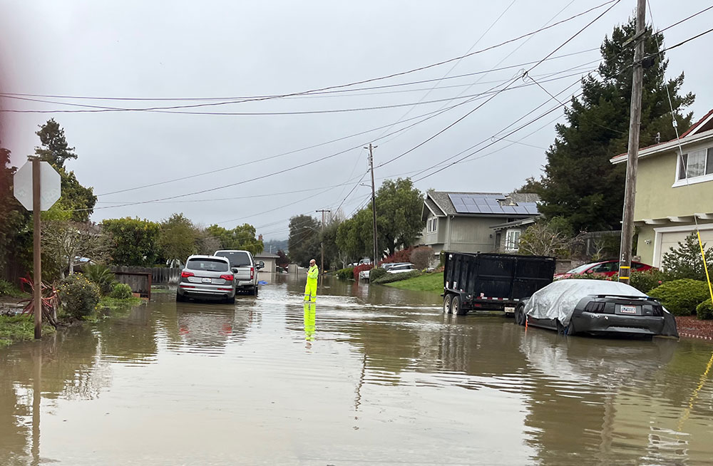 Red Cross volunteer in protective gear standing in waters on flooded residential street.