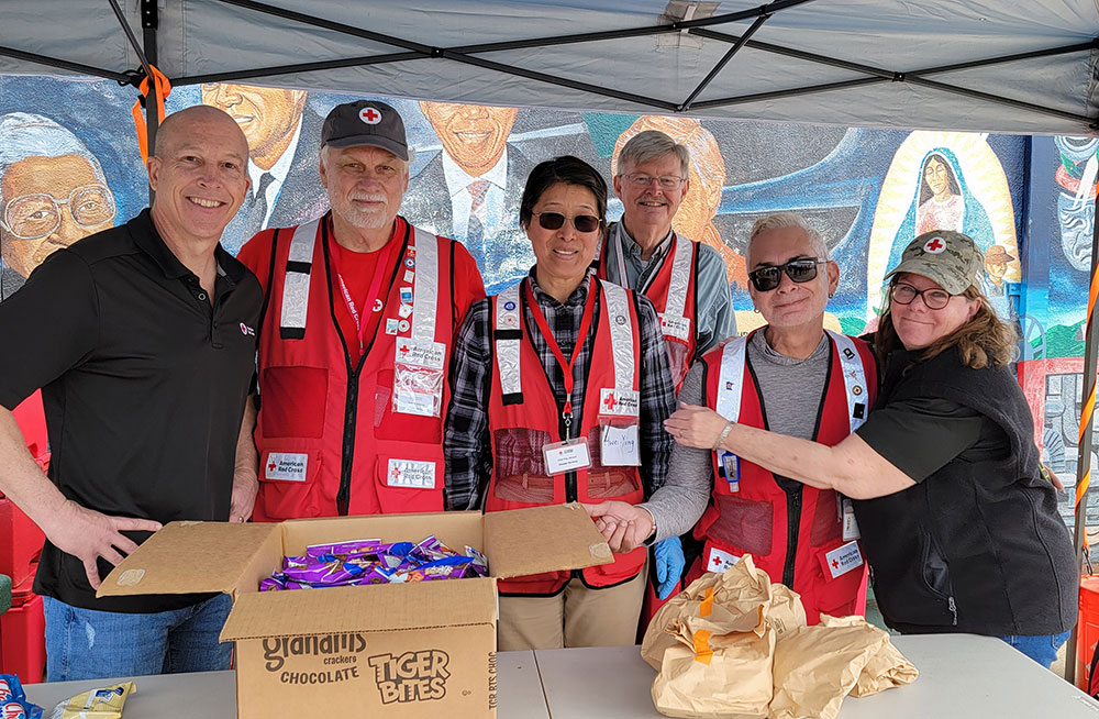Red Cross volunteers under canopy with snacks on a table.