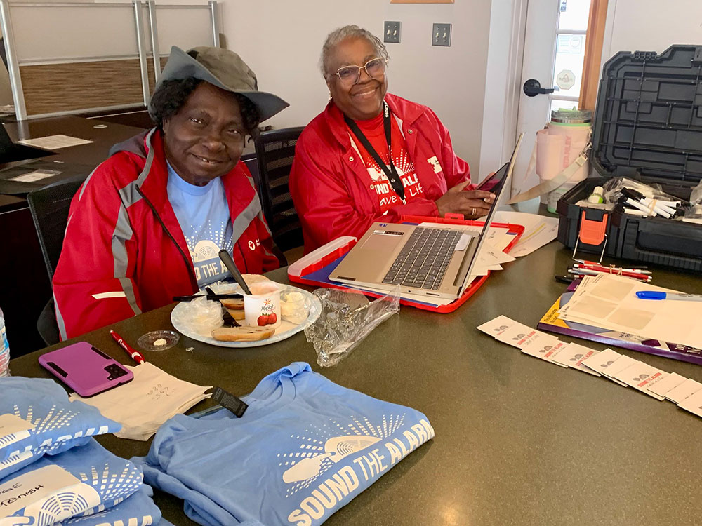 Red Cross Volunteers Alzinia Pailin and Vanessa Brown smiling in red jackets sit at a table with a laptop and papers, surrounded by kits and "Sound the Alarm" T-shirts.