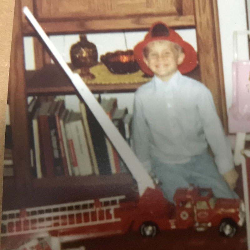 Brian Oftedal as a child wearing firefighter hat with toy fire truck