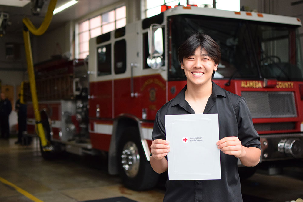 Edward Kuan holding the Red Cross Certificate of Extraordinary Personal Action in front of a fire engine.