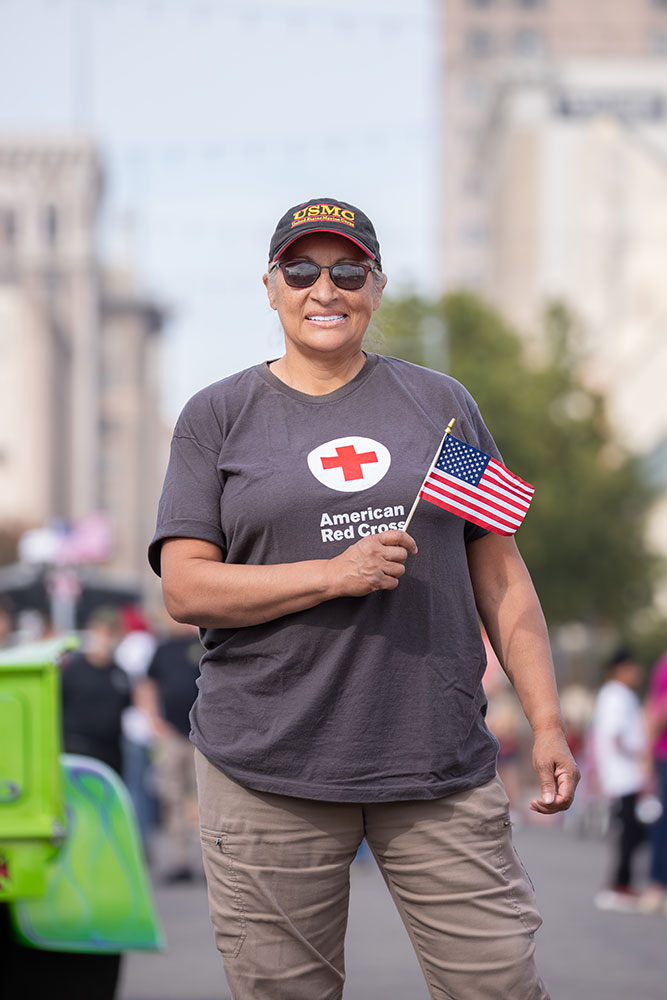 Susan Lugo wearing a hat, sunglasses, a Red Cross shirt and holding a small American flag.
