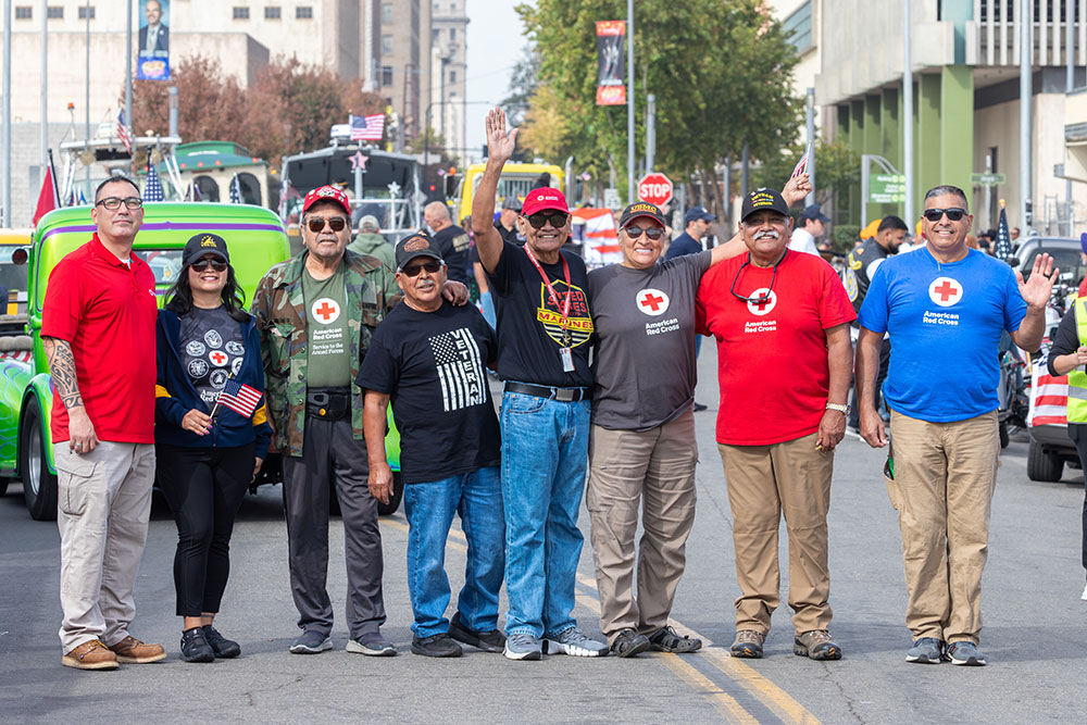 Susan Lugo wearing a hat, sunglasses and a Red Cross shirt stands next to seven veterans during an event on the city street.