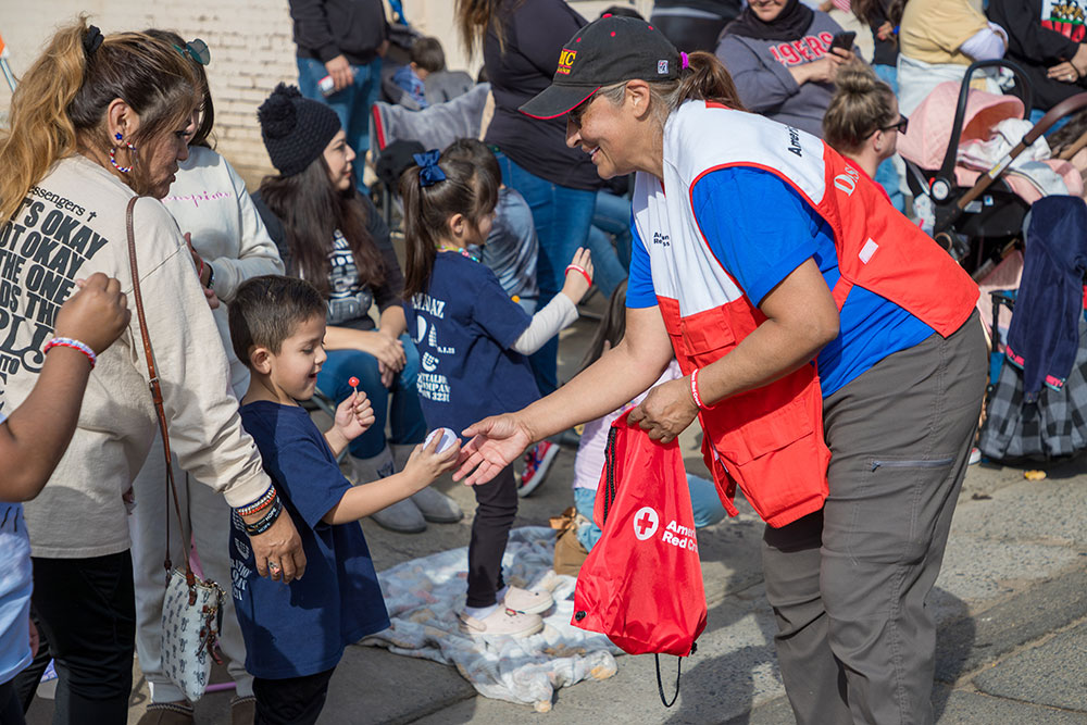 Susan Lugo wearing a hat and Red Cross vest hands a small gift to a child at an event on the street.