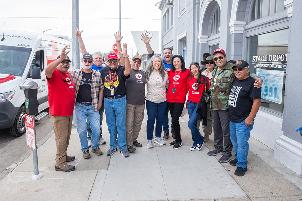 Susan Lugo wearing a hat, sunglasses and a Red Cross shirt stands next to veterans and Red Cross volunteers on a sidewalk at a Red Cross event.