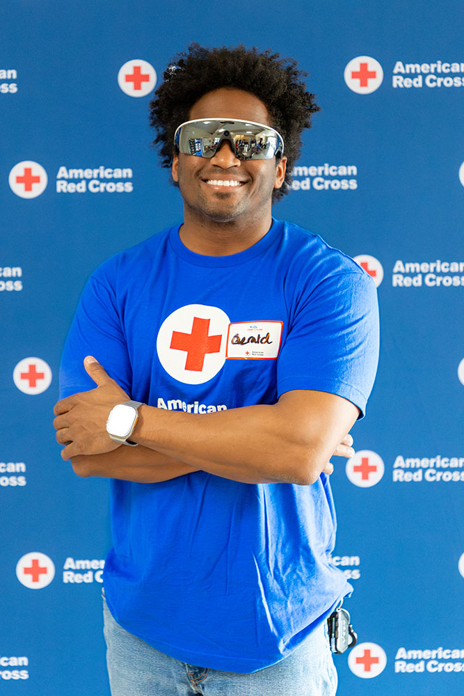 Gerald Golstein wearing sunglasses and a Red Cross shirts stands smiling with his arms crossed in front of a Red Cross logo backdrop.