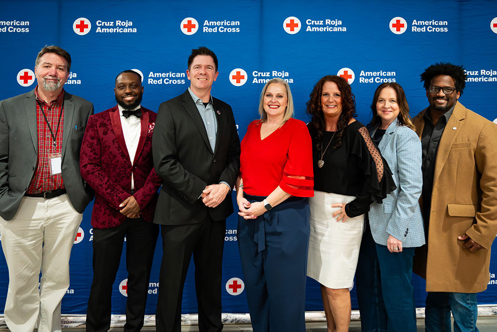 Gerald Golstein and six other people stand smiling in front of a Red Cross backdrop at a Red Cross event..